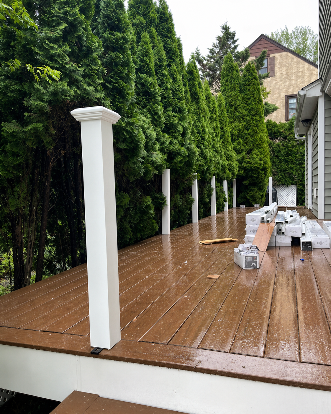 Wooden deck under construction beside tall green hedges, with white posts and building materials stacked on it