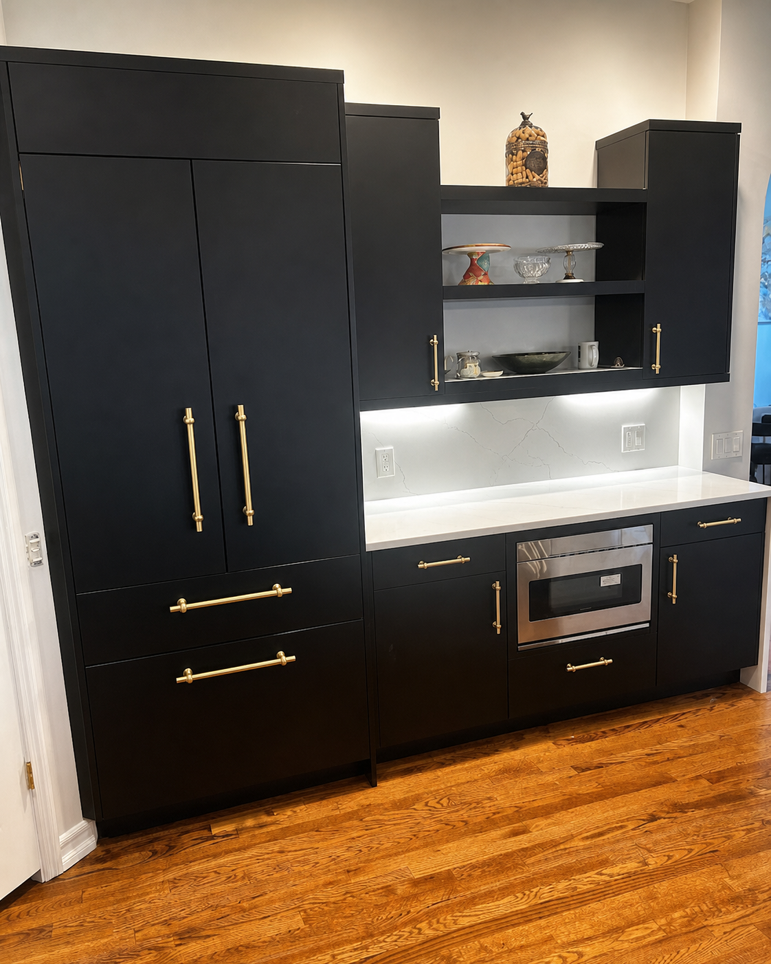 Black kitchen cabinets, built-in oven, and white countertop with shelves against a wood floor.
