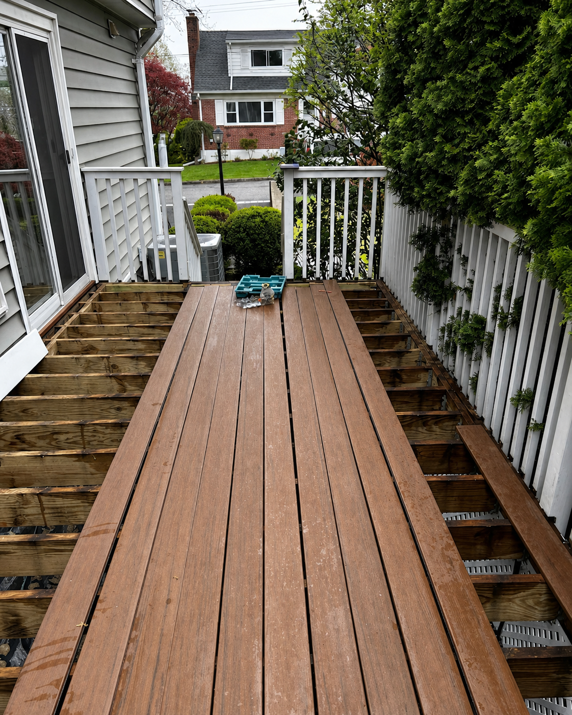 Narrow wooden deck walkway between white railings, leading to a yard and house beyond.