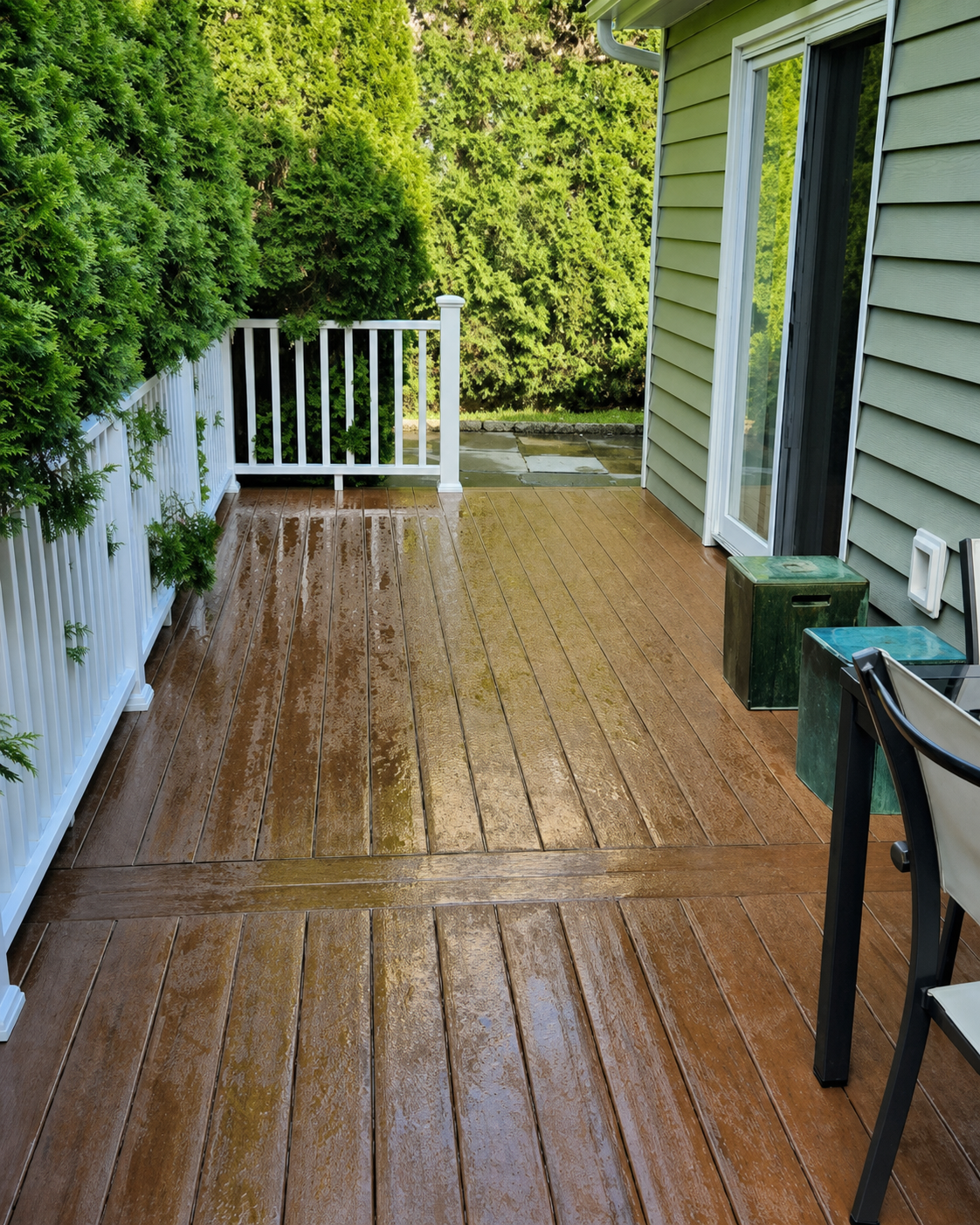 Wet wooden deck beside a house, with white railing, green hedges, and a glass door.