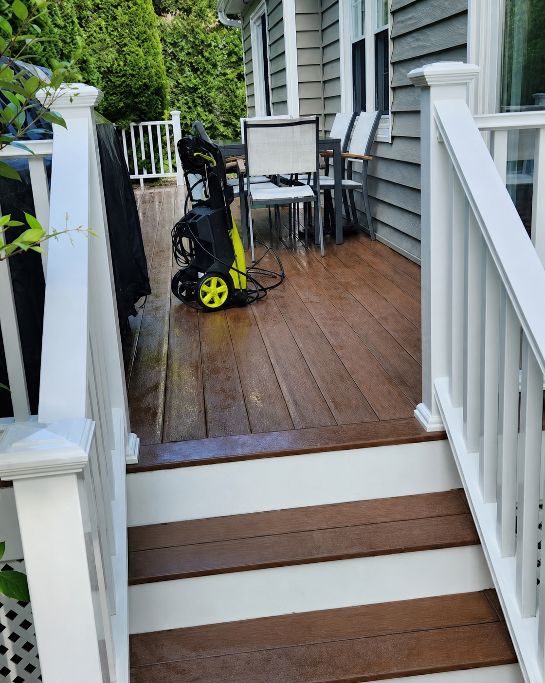 Wet wooden deck with patio table, chair, and pressure washer beside white railings and house siding