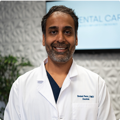 Man in white coat, smiling, with gray beard; in a dental office.