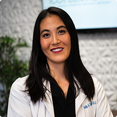 Woman in a white coat smiling, looking at the camera. Neutral background, indoors.