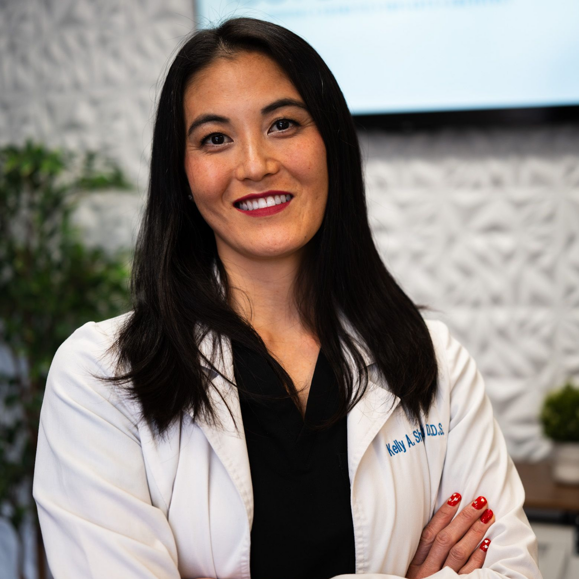 A professional with long dark hair, wearing a white lab coat over a black shirt, posing with arms crossed in an office.