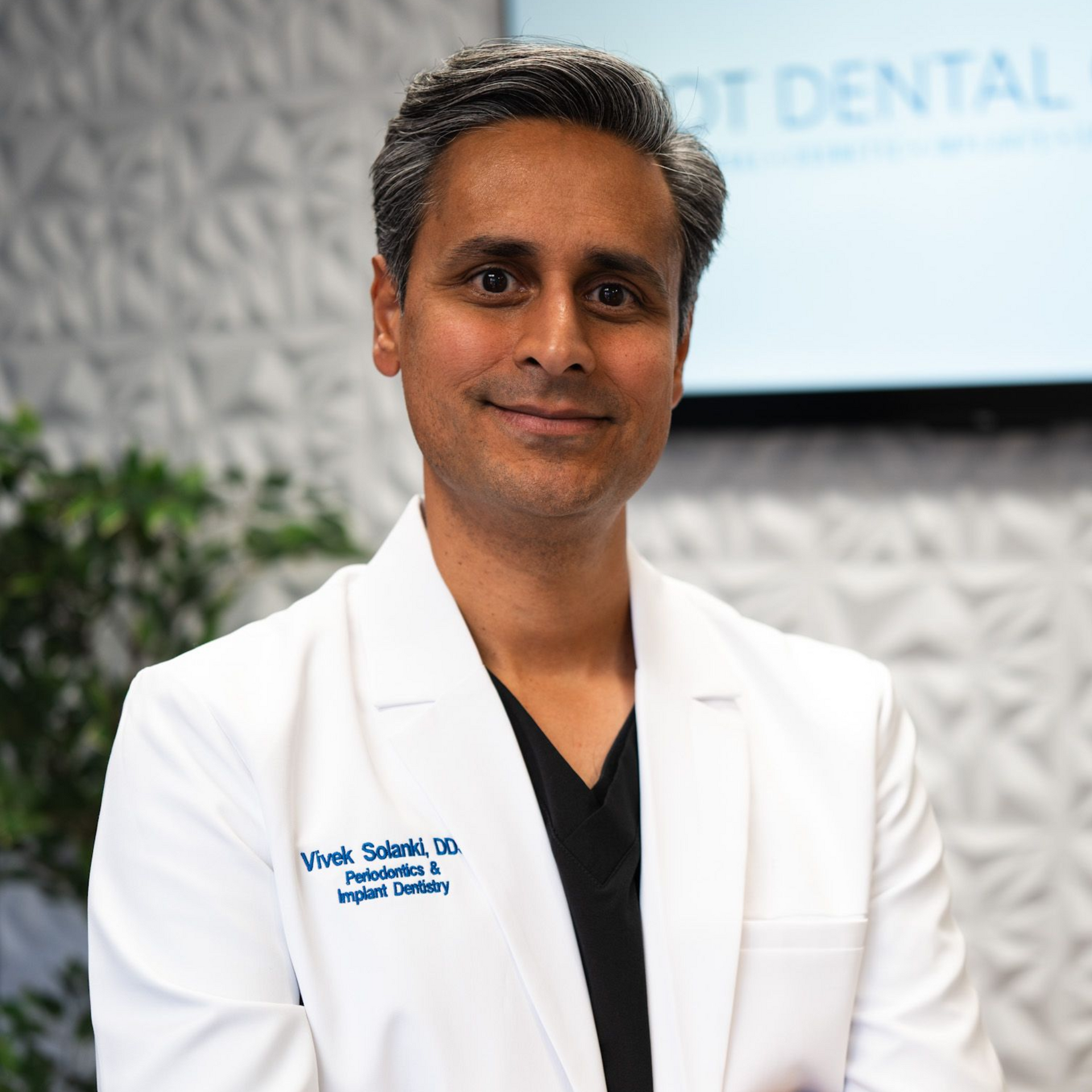Professional headshot of a dentist in a white coat and black scrubs, standing in an office with a textured wall.