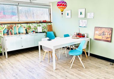 Een lichte klaslokaal met een witte tafel, blauwe stoelen, kasten en kleurrijke wanddecoraties.