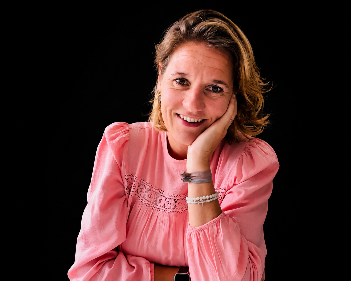 Een lachende vrouw in een roze blouse, die haar kin op haar hand laat rusten, tegen een zwarte achtergrond.