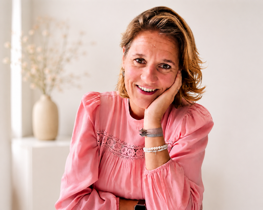 Een lachende vrouw in een roze blouse rust met haar kin op haar hand in een lichte kamer.