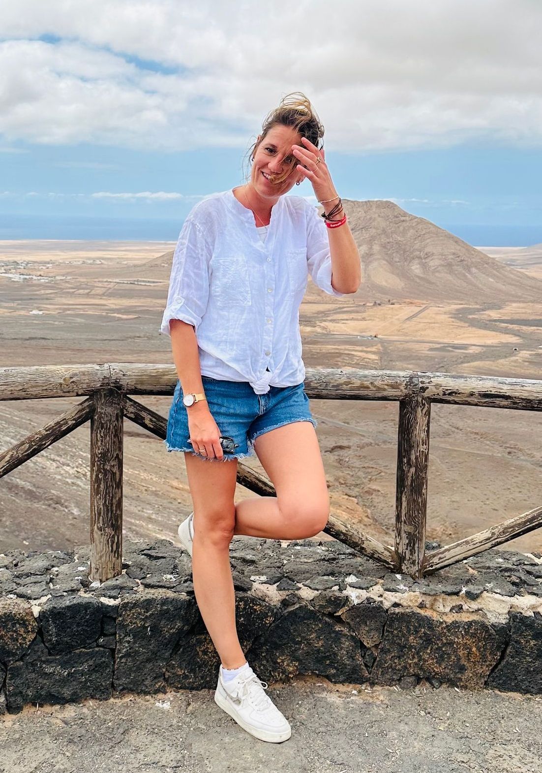 Vrouw in witte blouse en spijkerbroek poseert bij een stenen muur met bergen en een bewolkte hemel op de achtergrond.
