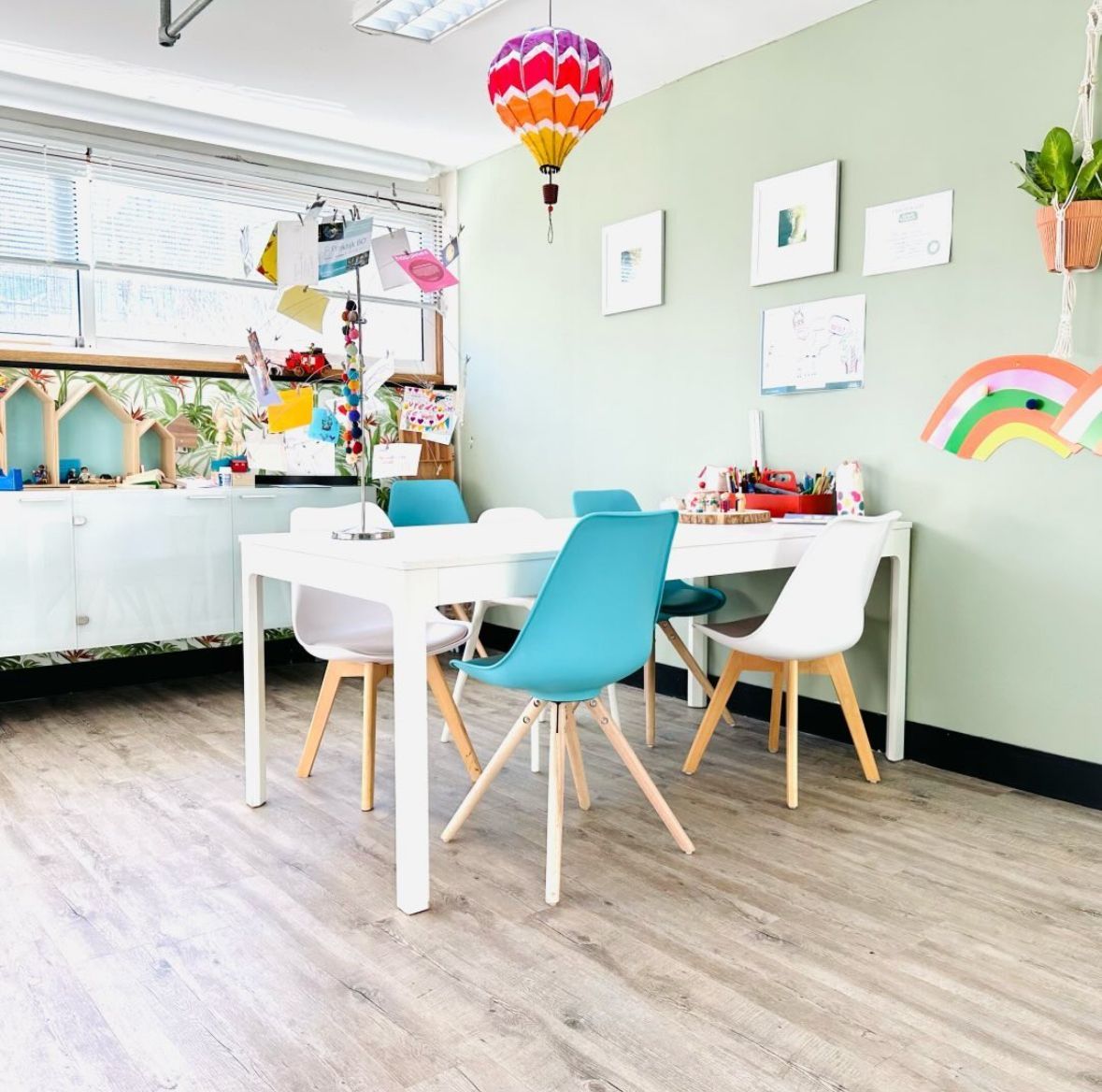 Een vrolijke kinderkamer met een witte tafel, een turquoise stoel, kleurrijke muurdecoratie en regenboogaccenten.