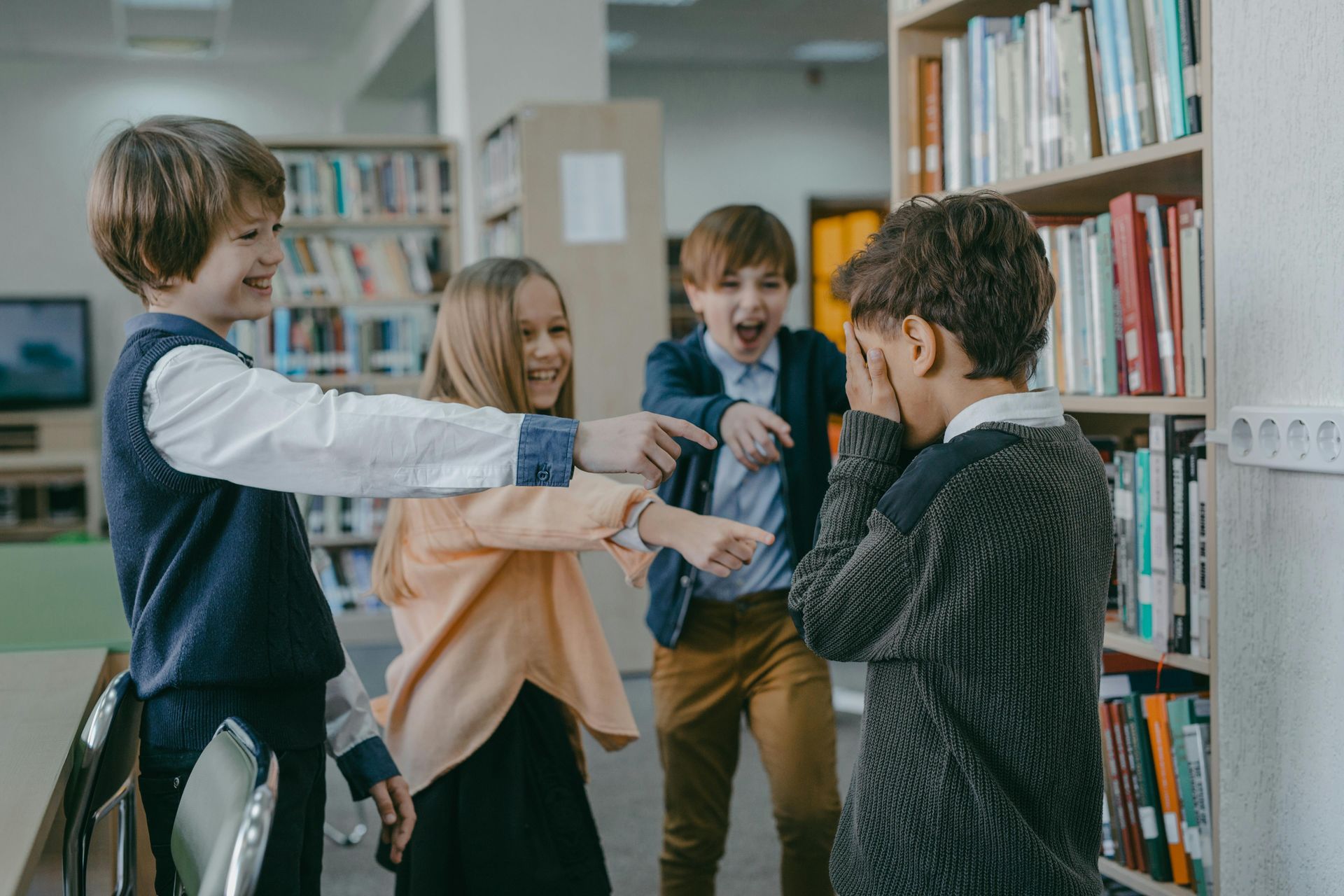 Kinderen lachen en wijzen naar een ander kind dat zijn gezicht bedekt. ​​In een bibliotheek.