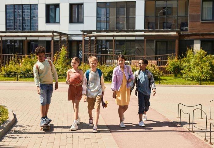 Vijf studenten lopen samen buiten een modern campusgebouw op een zonnige dag.