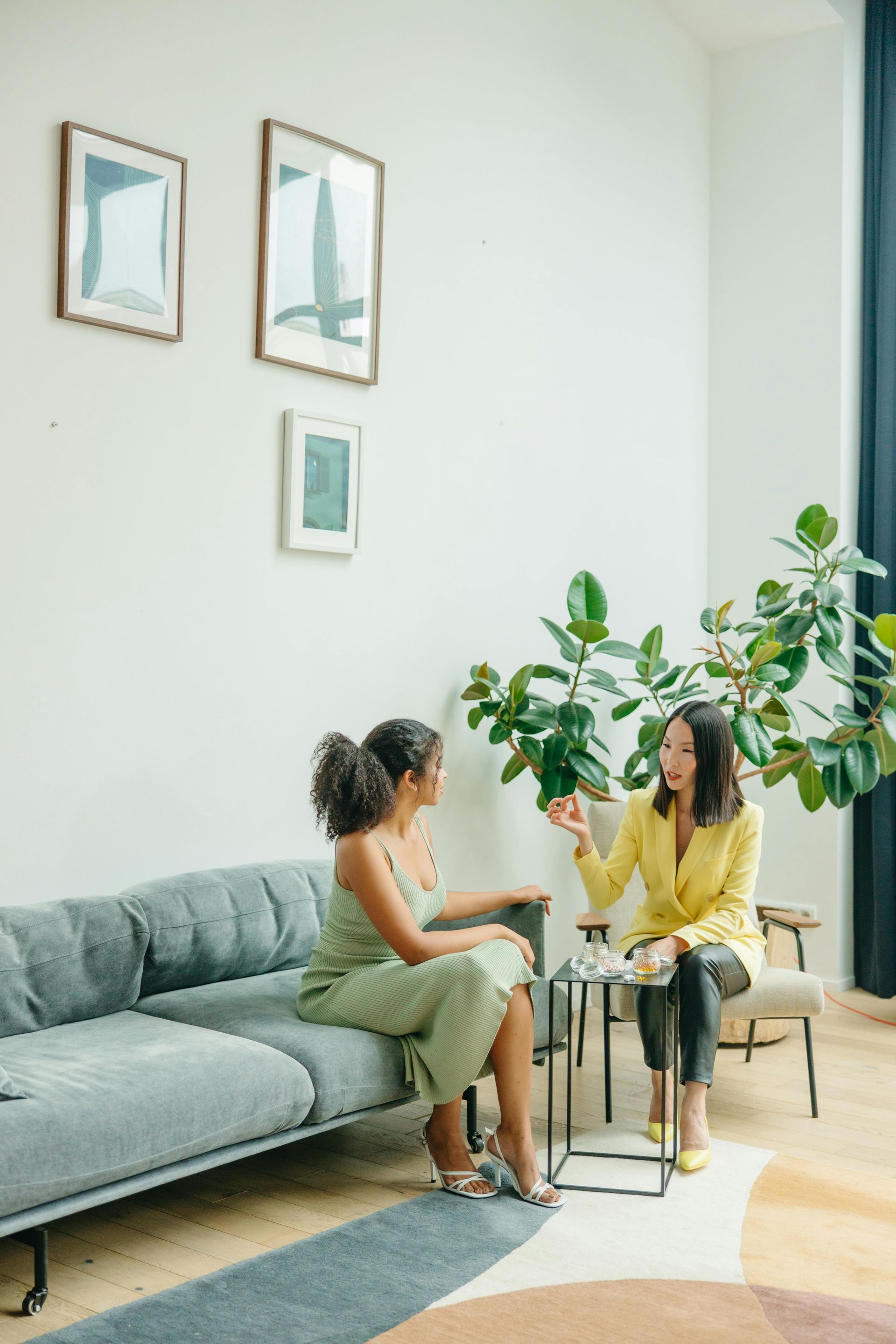 Twee mensen praten aan een kleine tafel in een lichte woonkamer met een grijze bank en een grote groene plant.