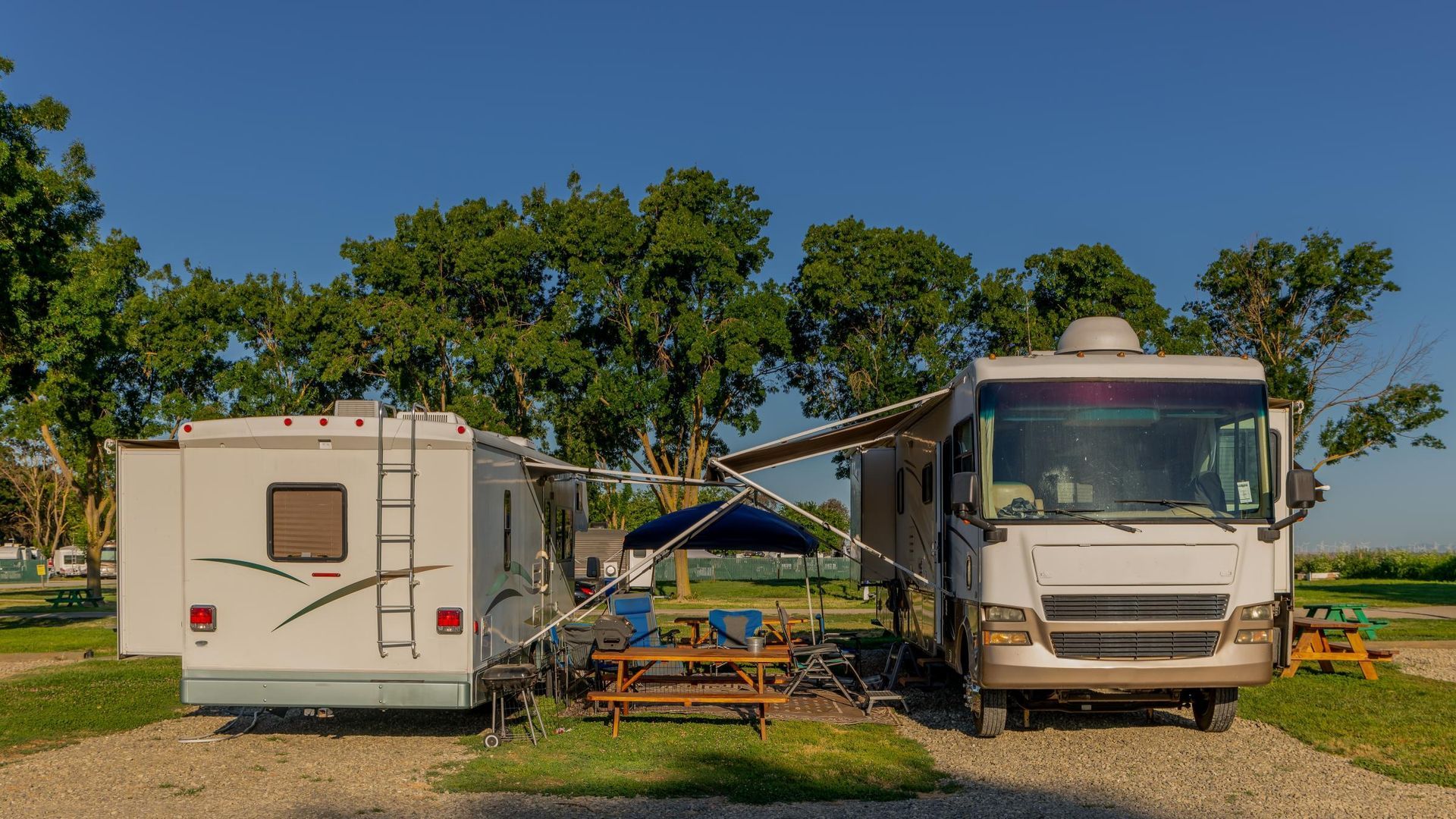 Two RVs parked side-by-side at a campsite on a sunny day. Blue sky, green trees, and picnic table.