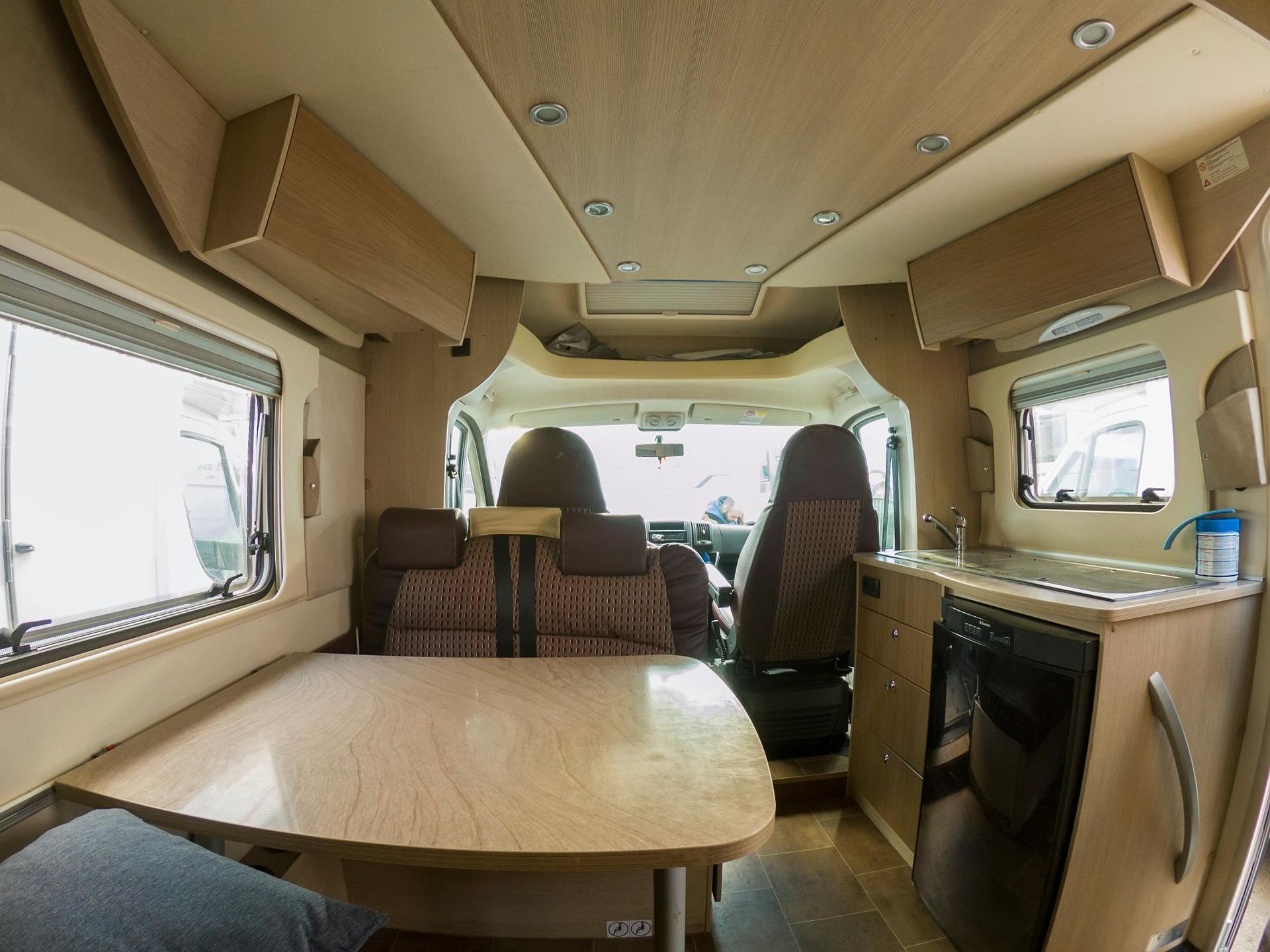 Interior of a motorhome with a table, seating, kitchen, and driver's area. Light wood and beige tones.