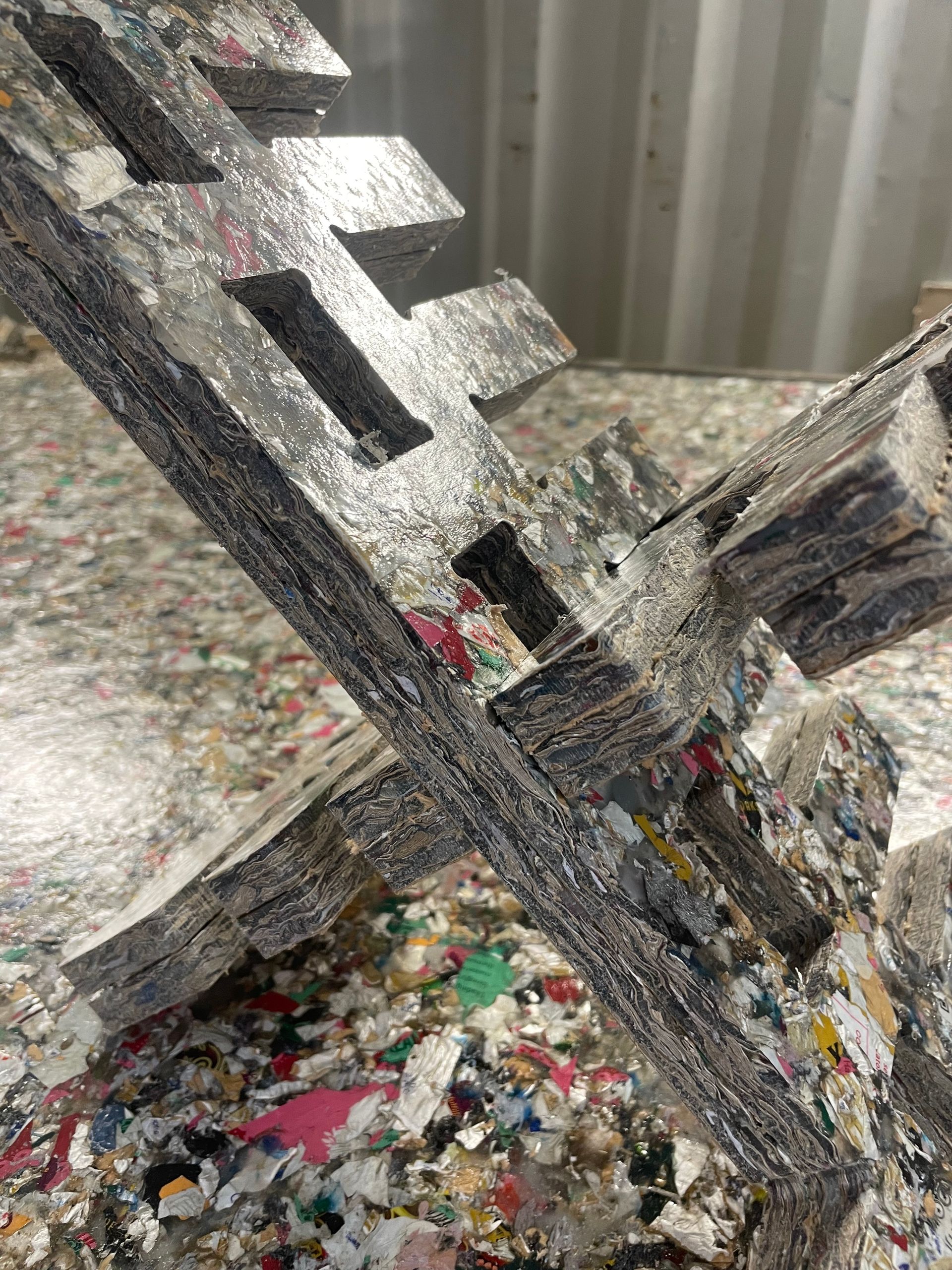 saveBOARD. Close-up of a dark, cross-shaped object with cut-out rectangles, amidst a pile of colorful plastic flakes.