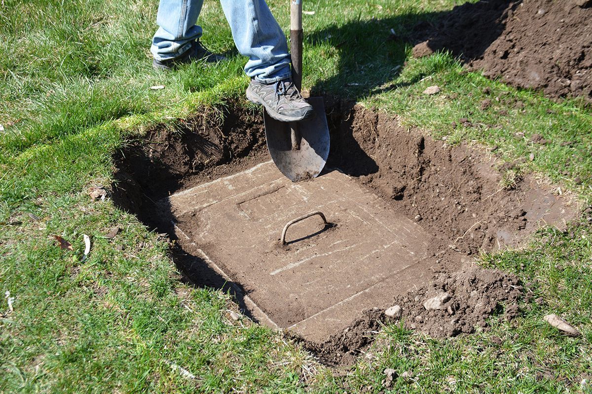 Person digging around a concrete underground utility access cover in grass, revealing the lid with a handle.
