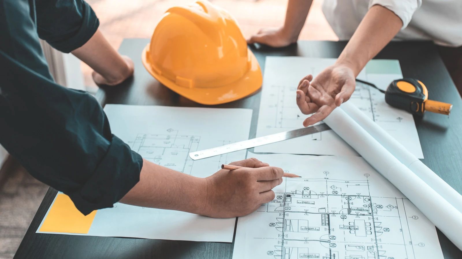 Two people collaborating on blueprints, with a yellow hard hat on a table.