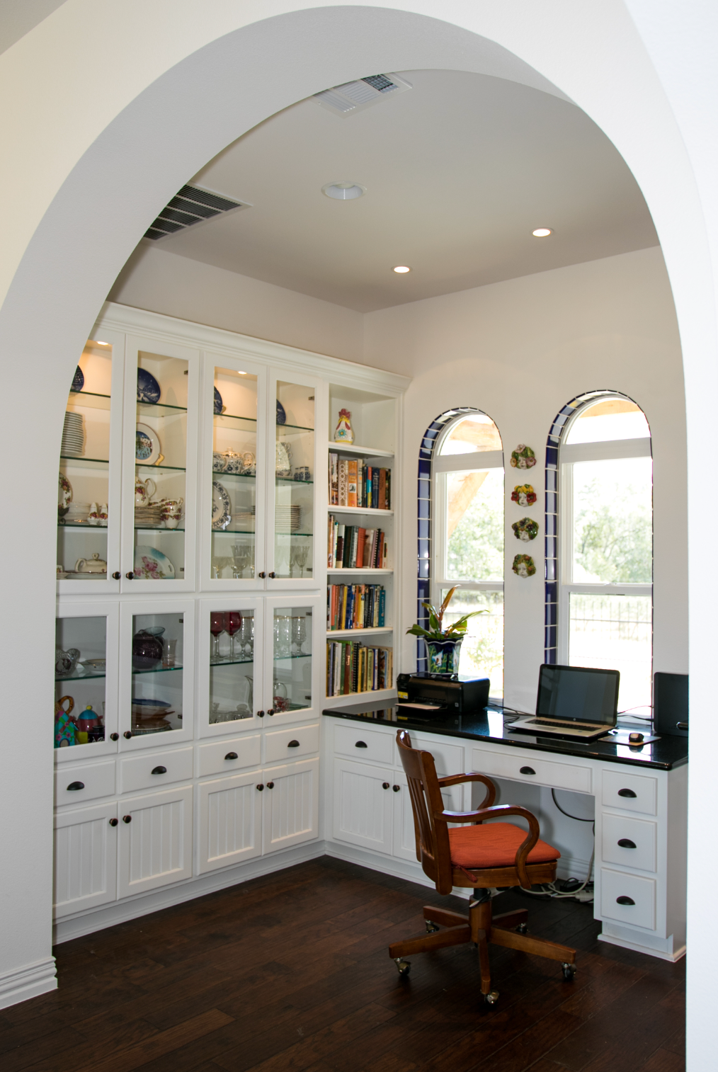 A home office with a desk , chair , bookshelves and arched windows.