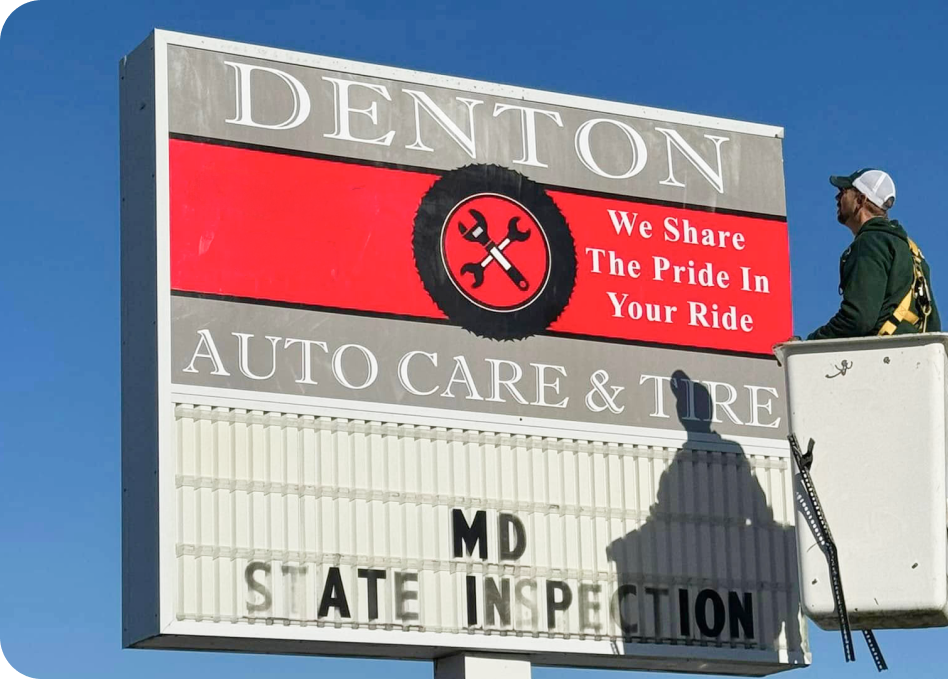 Denton Auto Care & Tire roadside sign with red “We Share The Pride In Your Ride” panel and a worker on top | Denton Auto Care And Tire