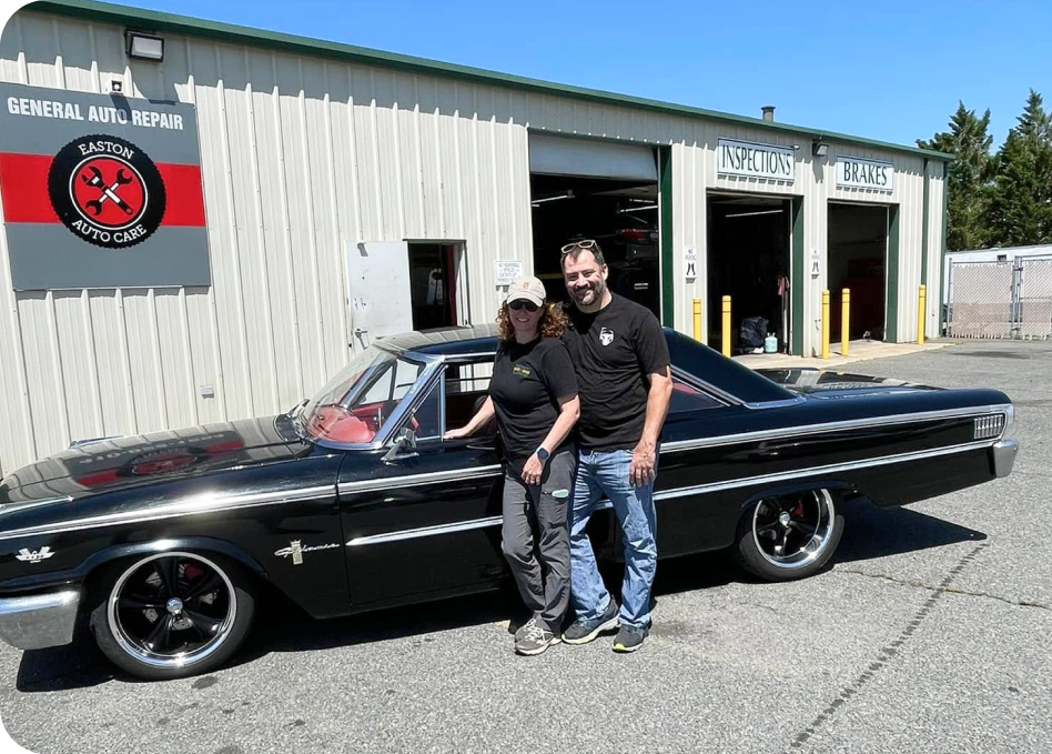 Two people posing beside a black classic car outside a garage on a sunny day | Denton Auto Care And Tire