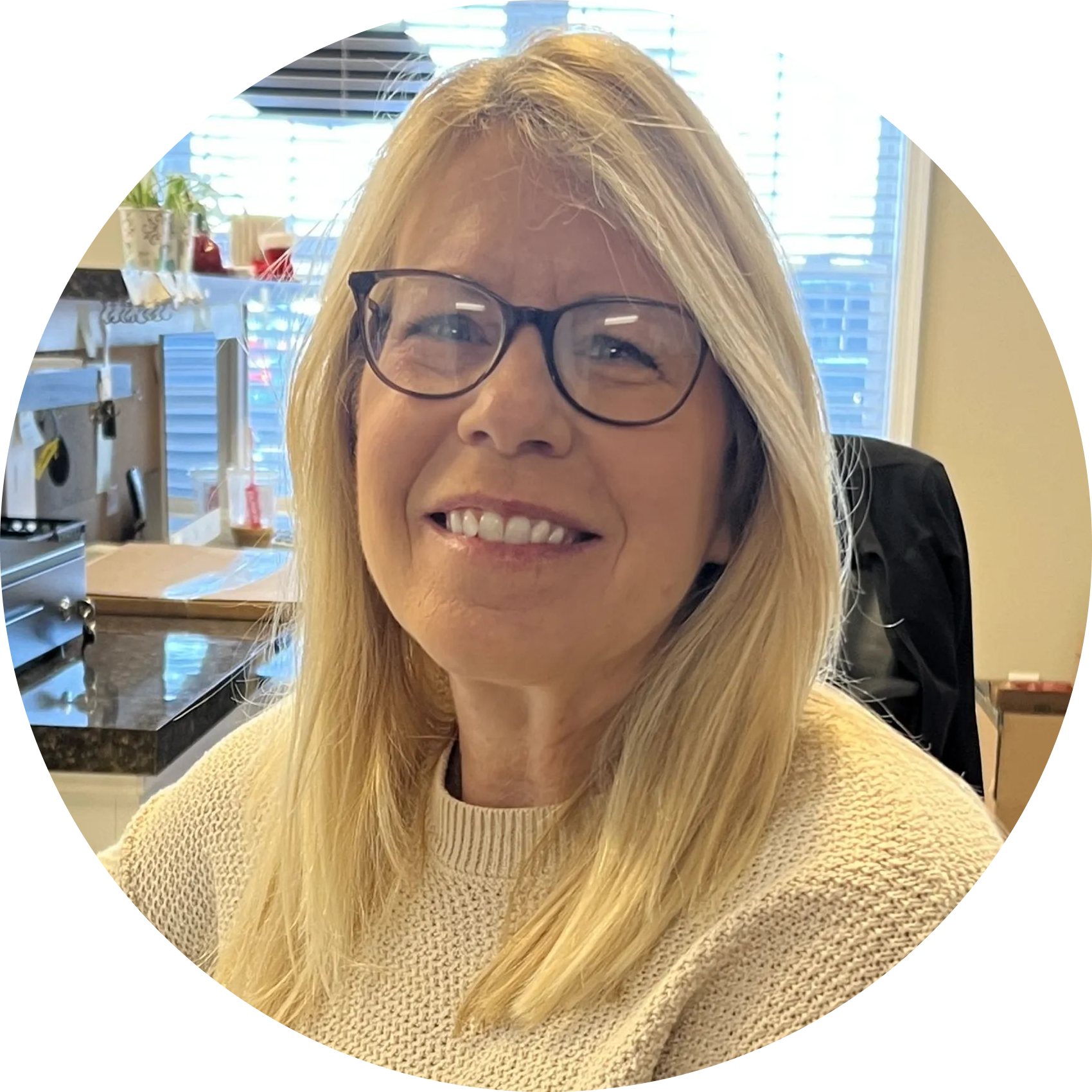 Smiling woman with glasses in a cream sweater, seated in an office with a window and city view. | Denton Auto Care And Tire