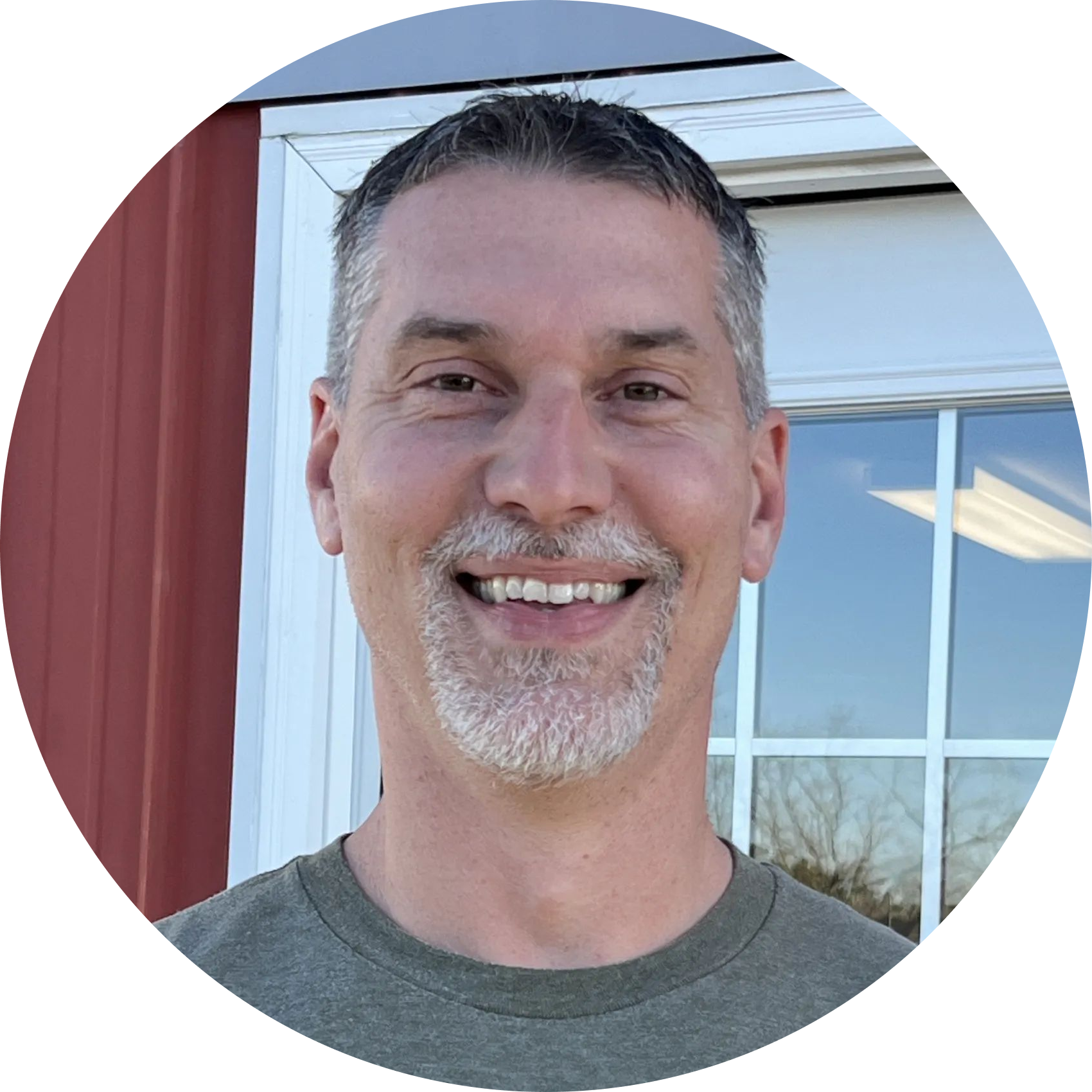Smiling person in a gray shirt standing outdoors beside a red wall and window. | Denton Auto Care And Tire