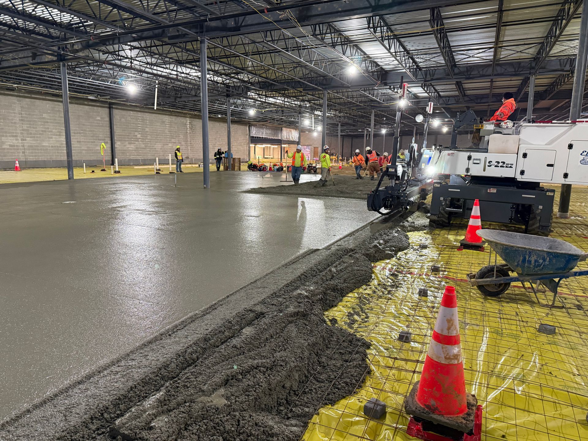 Workers paving a large indoor warehouse floor with fresh gray concrete and orange cones nearby.