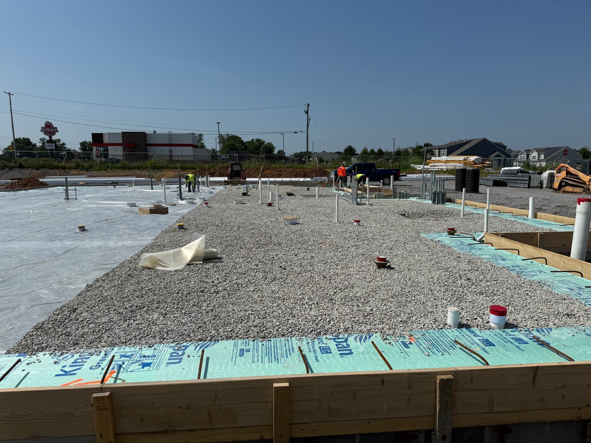 Construction site with gravel foundation, wooden framing, and workers under a clear blue sky