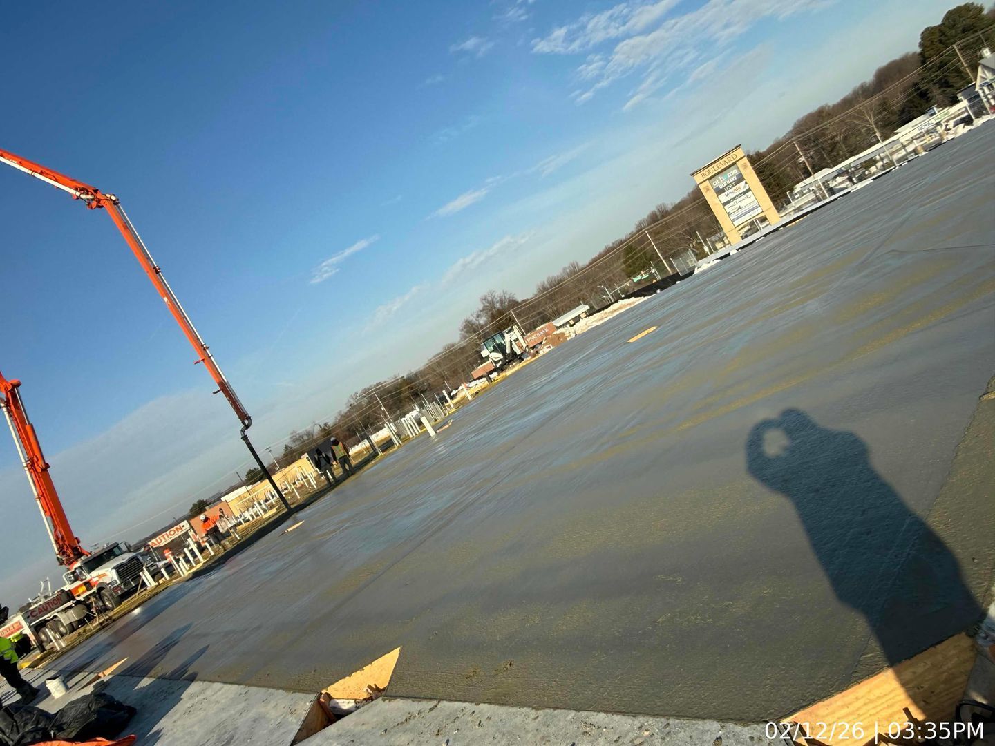 Tilted view of a concrete pour site with a red crane, wet slab, and buildings in the distance