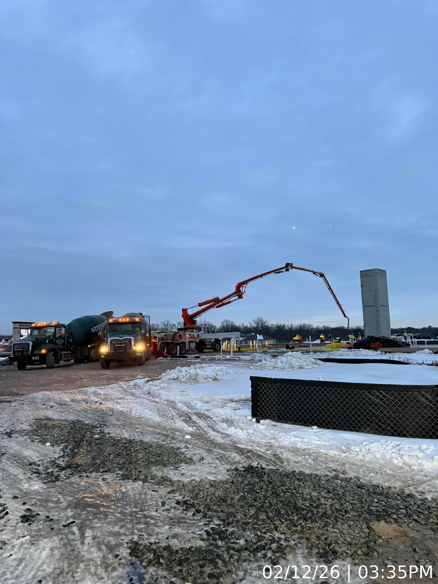 Construction trucks and a red concrete pump on a snowy site at dusk, with a tall building in the background