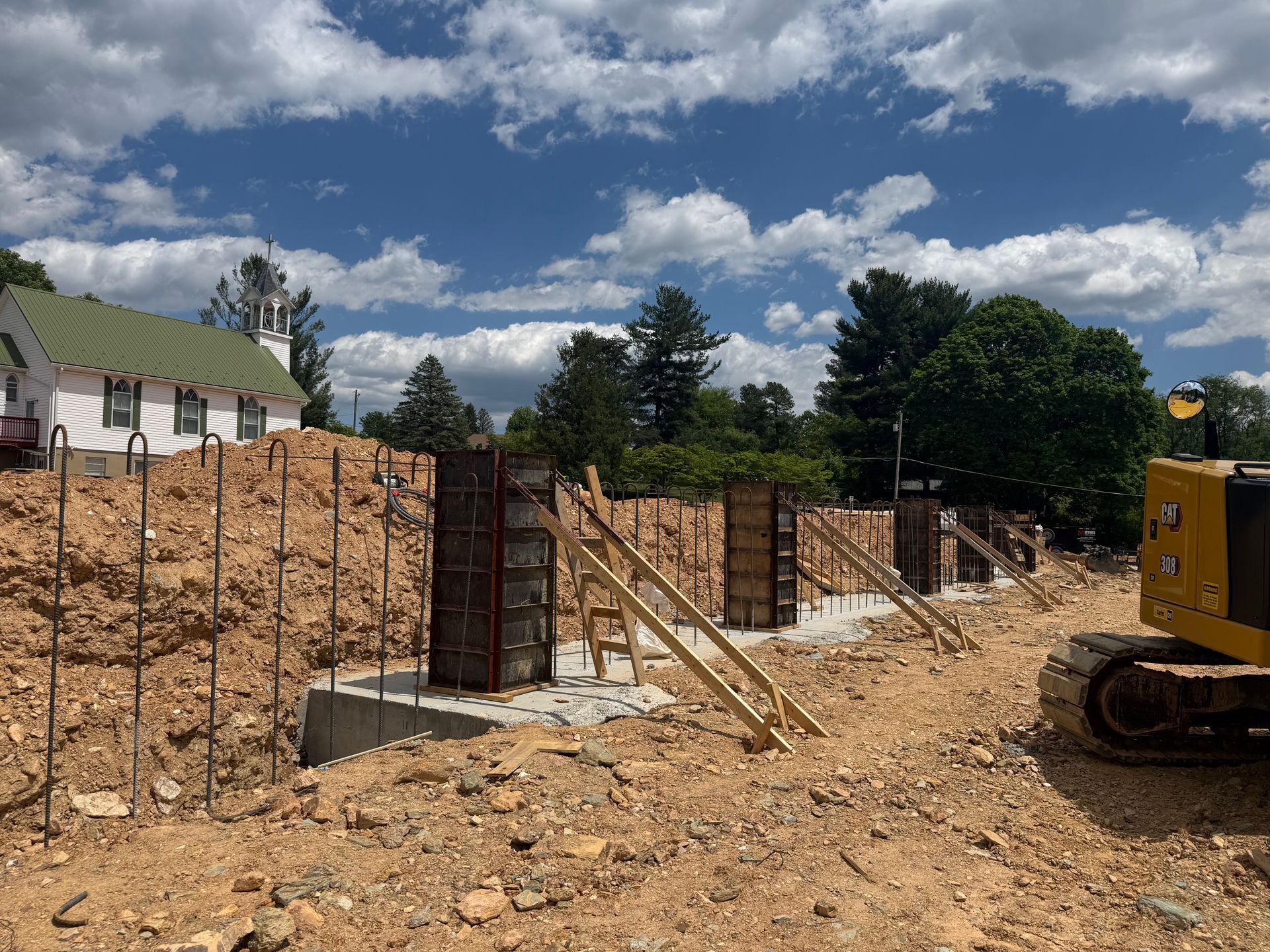 Construction site with concrete wall forms, dirt mound, and excavator under a cloudy sky