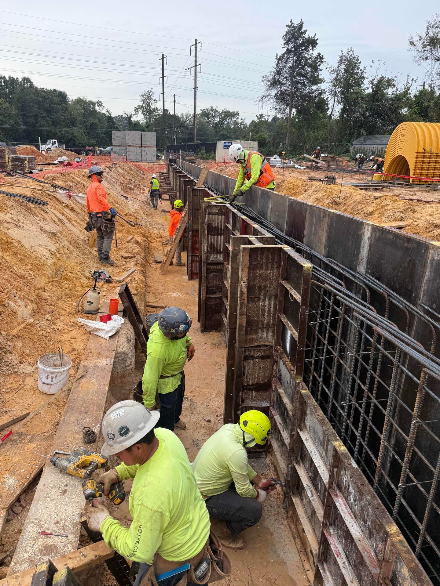 Construction workers in neon gear building a concrete trench wall at a muddy roadside site