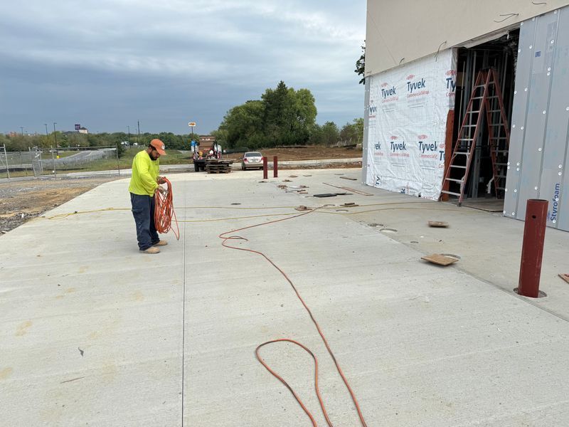 Worker in yellow vest unrolling cable on a concrete sidewalk beside a building under construction.