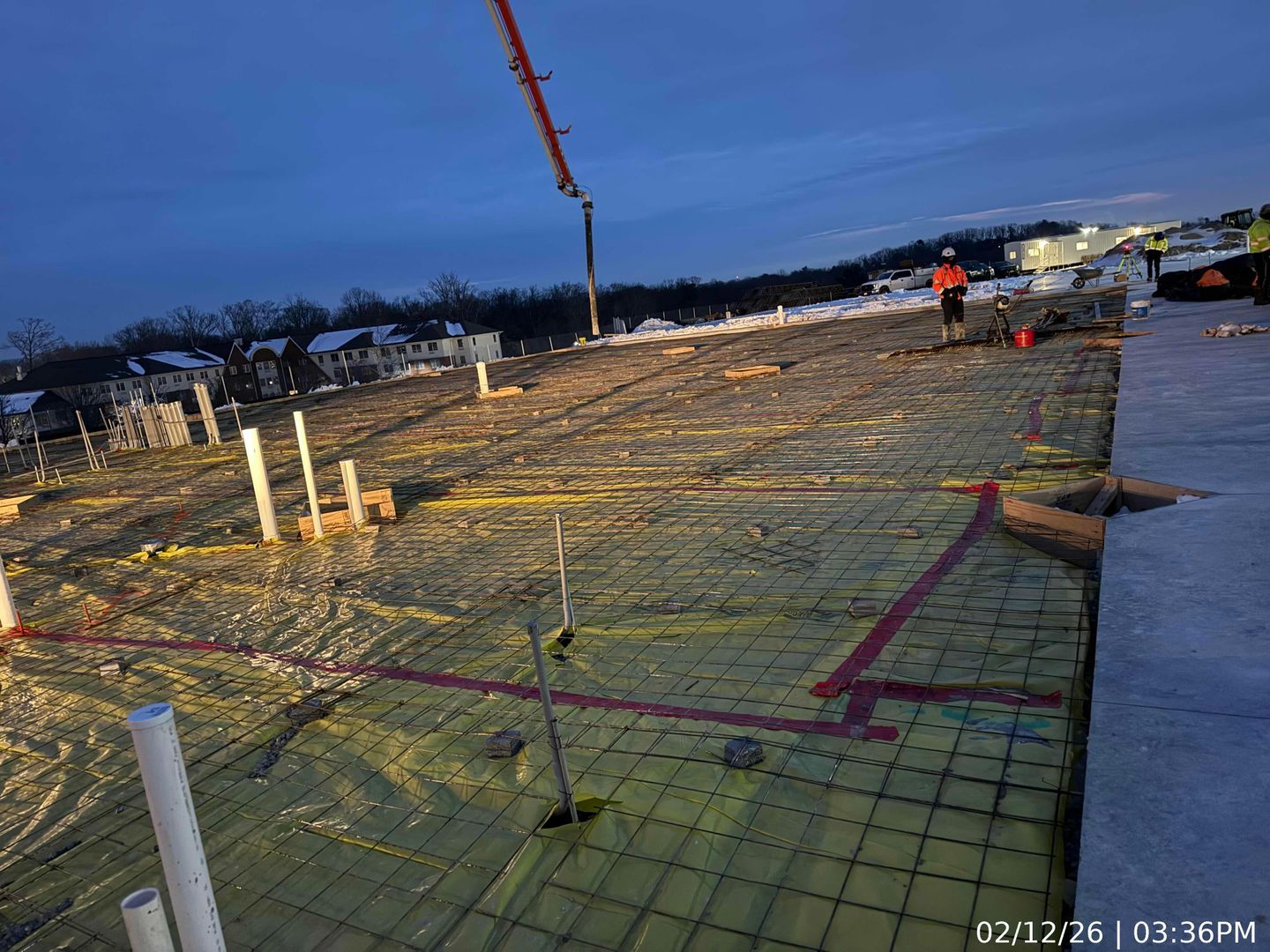 Rebar foundation slab under construction at dusk, with concrete forms, workers, and an excavator nearby.