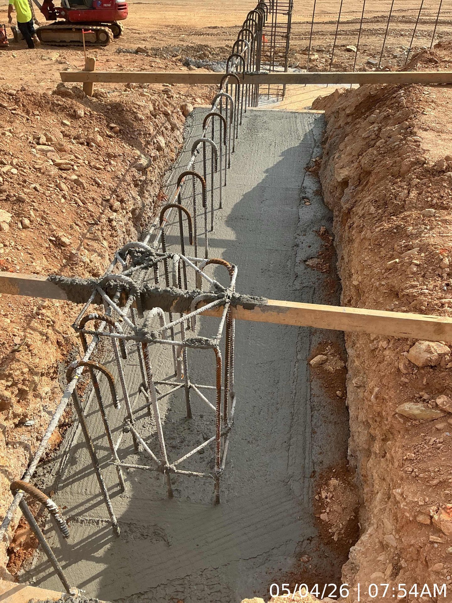 Concrete foundation trench with rebar framework and wooden supports at a construction site.