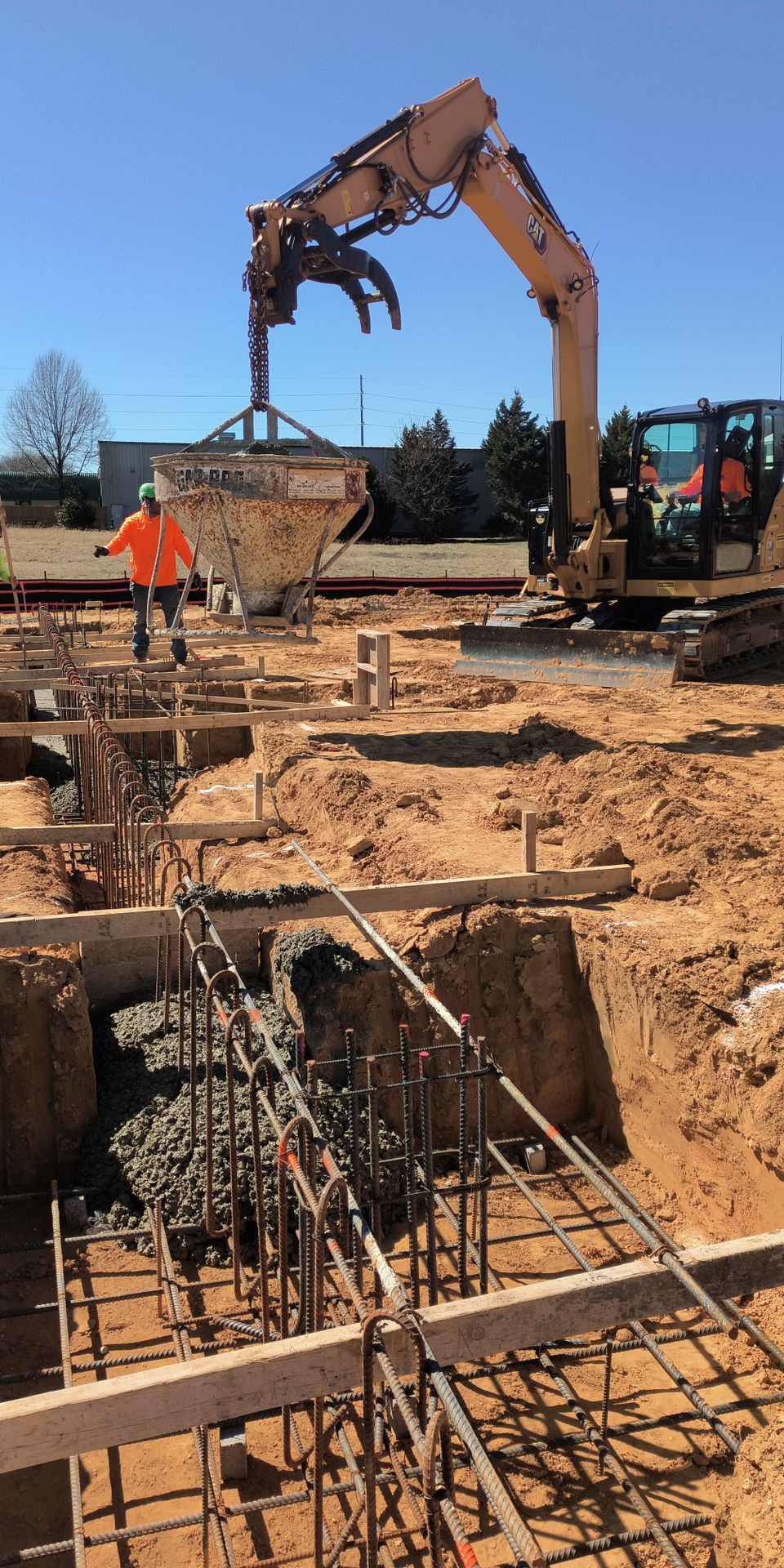 Excavator lifting a metal basket at a construction site with trenches and rebar in the foreground