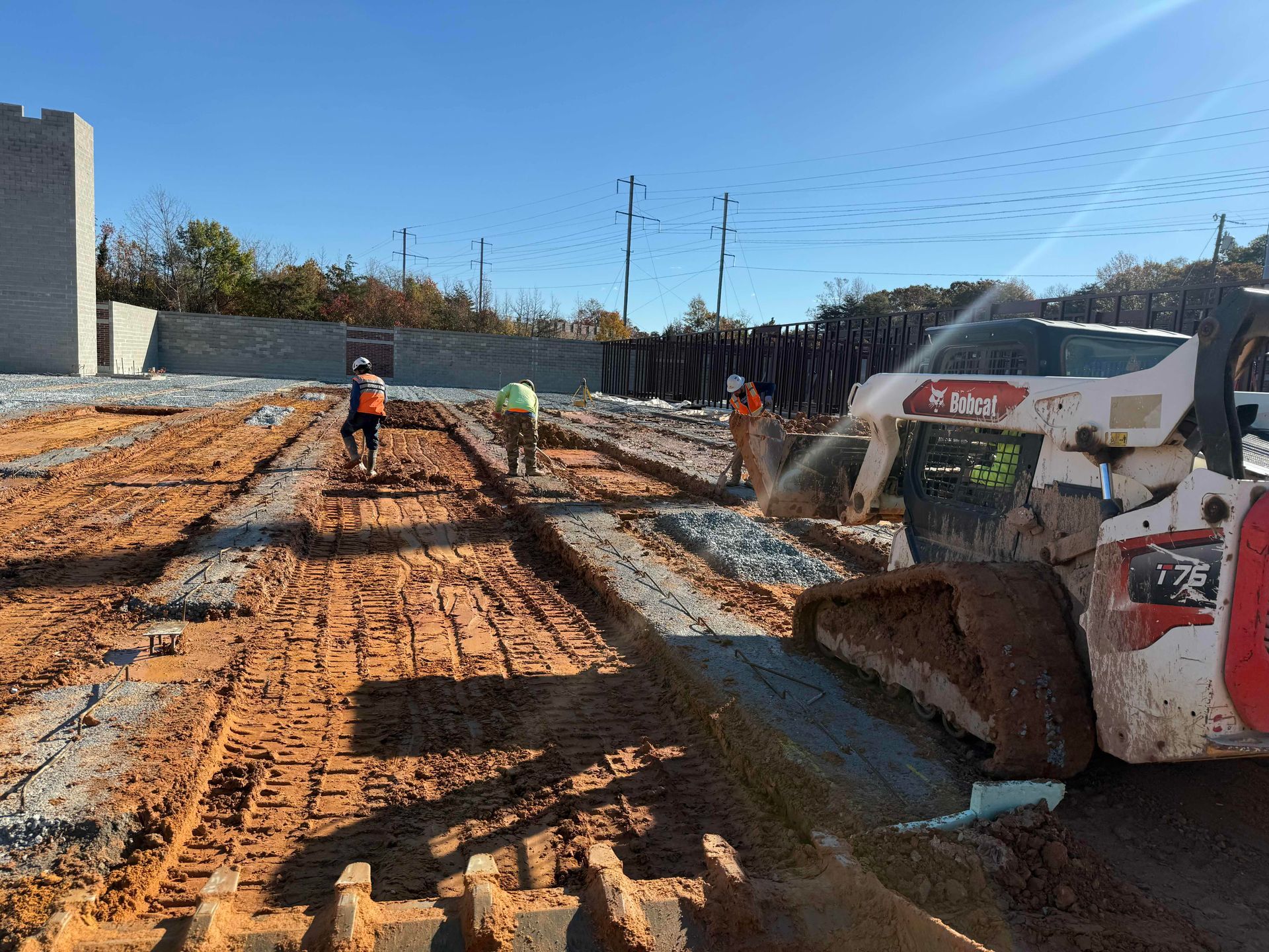 Construction site with workers leveling muddy ground beside a skid steer loader under a clear sky