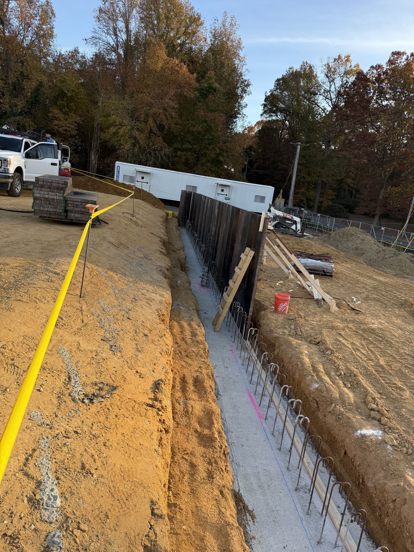 Construction trench beside a road with yellow cordon tape, steel retaining wall, and excavator nearby