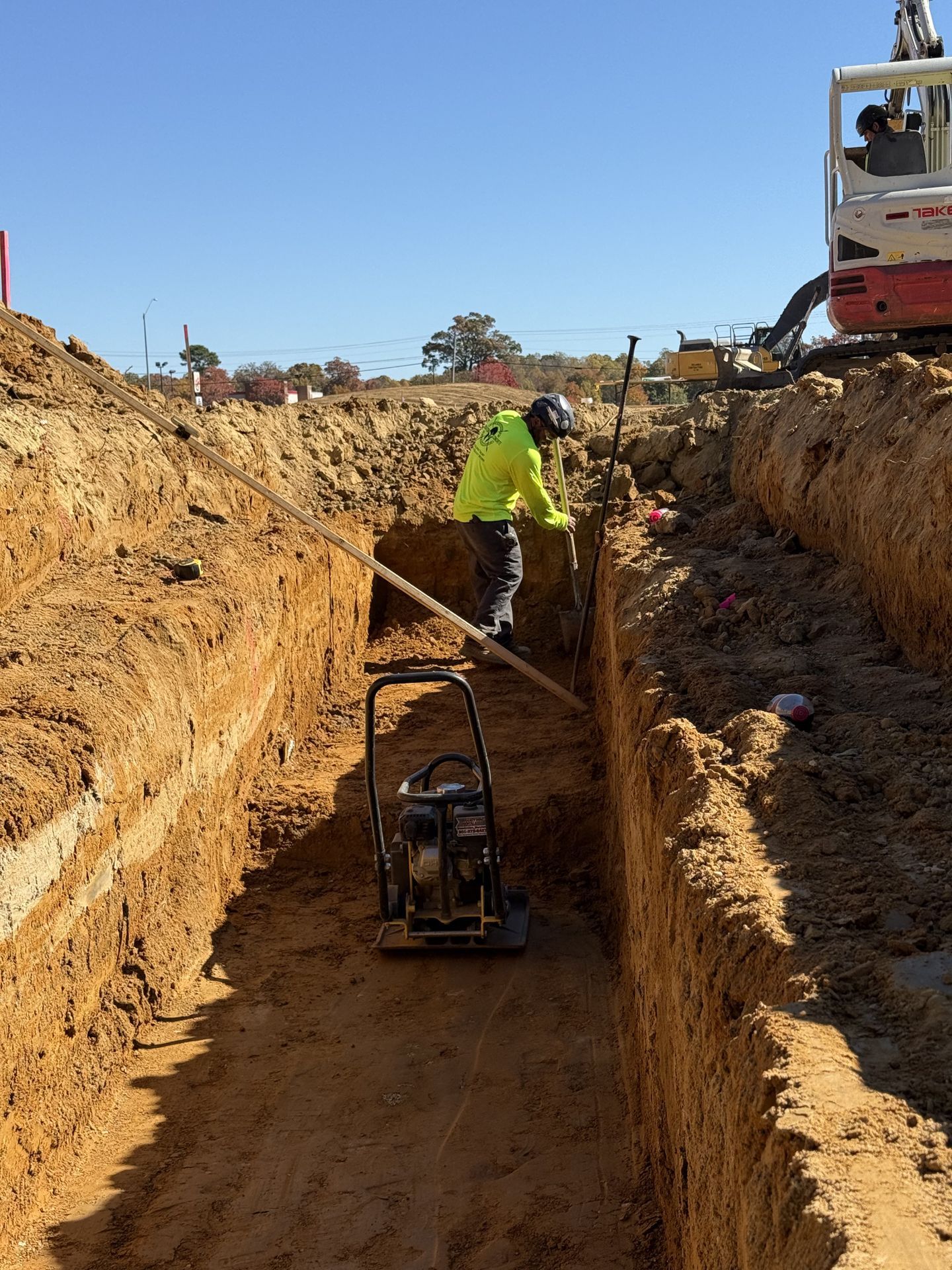 Construction workers in a deep trench operating a trench compactor beside an excavator
