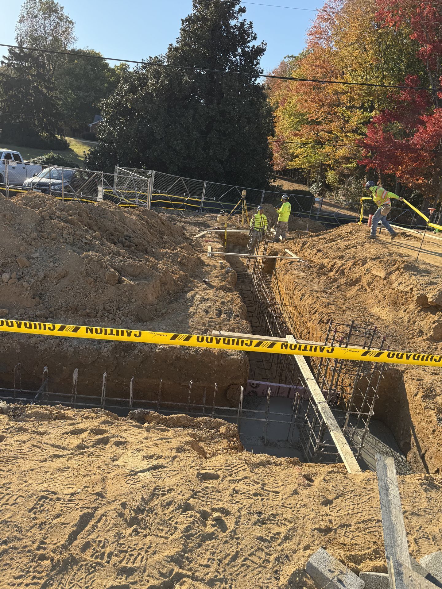 Construction trench with yellow caution tape, workers, and a wooden plank over sandy excavation outdoors