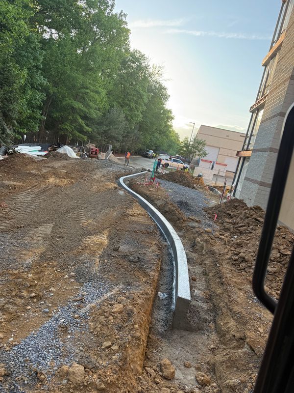 Construction site with curving white pipe laid along a dirt trench beside a building and trees