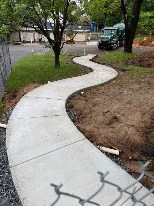 Curving concrete sidewalk through a wooded park, with a truck and work area in the background.