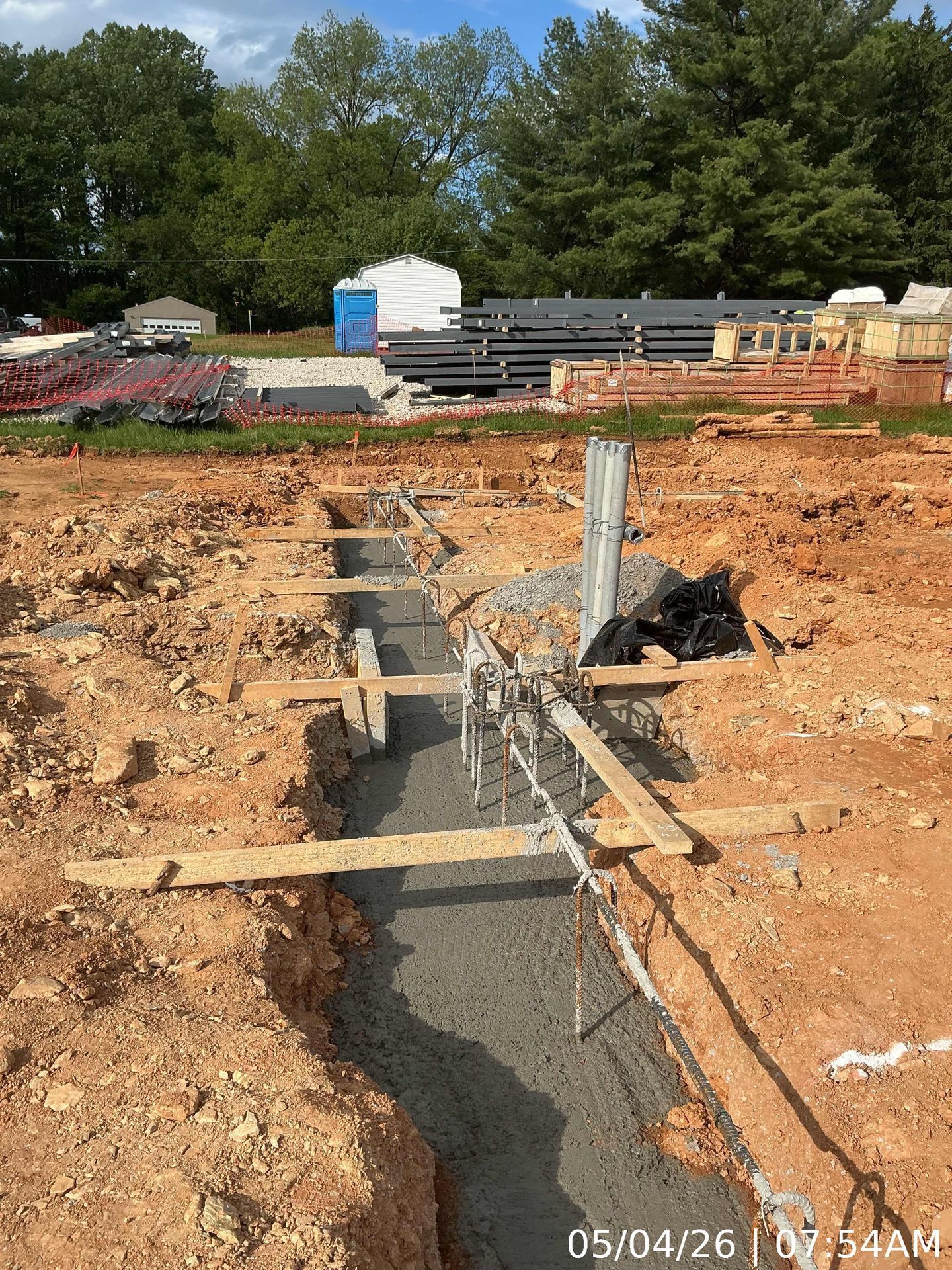 Foundation trench at a construction site with rebar and concrete forms in red clay soil, stacks of lumber in background