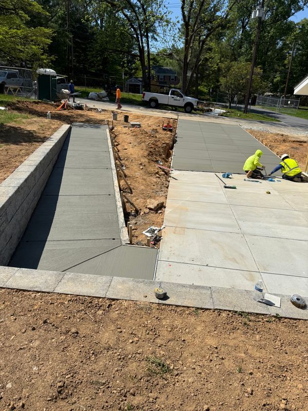 Workers installing concrete sidewalk ramp beside a road, with exposed dirt and curb edges.