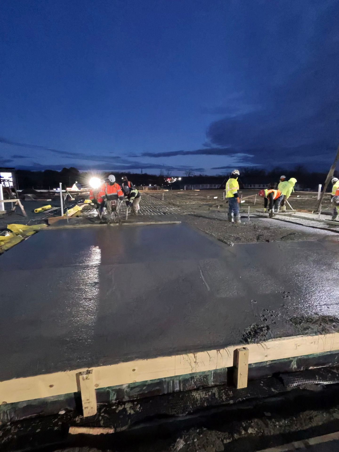 Workers pouring concrete on a roadway at dusk, with fresh wet concrete and construction lights.