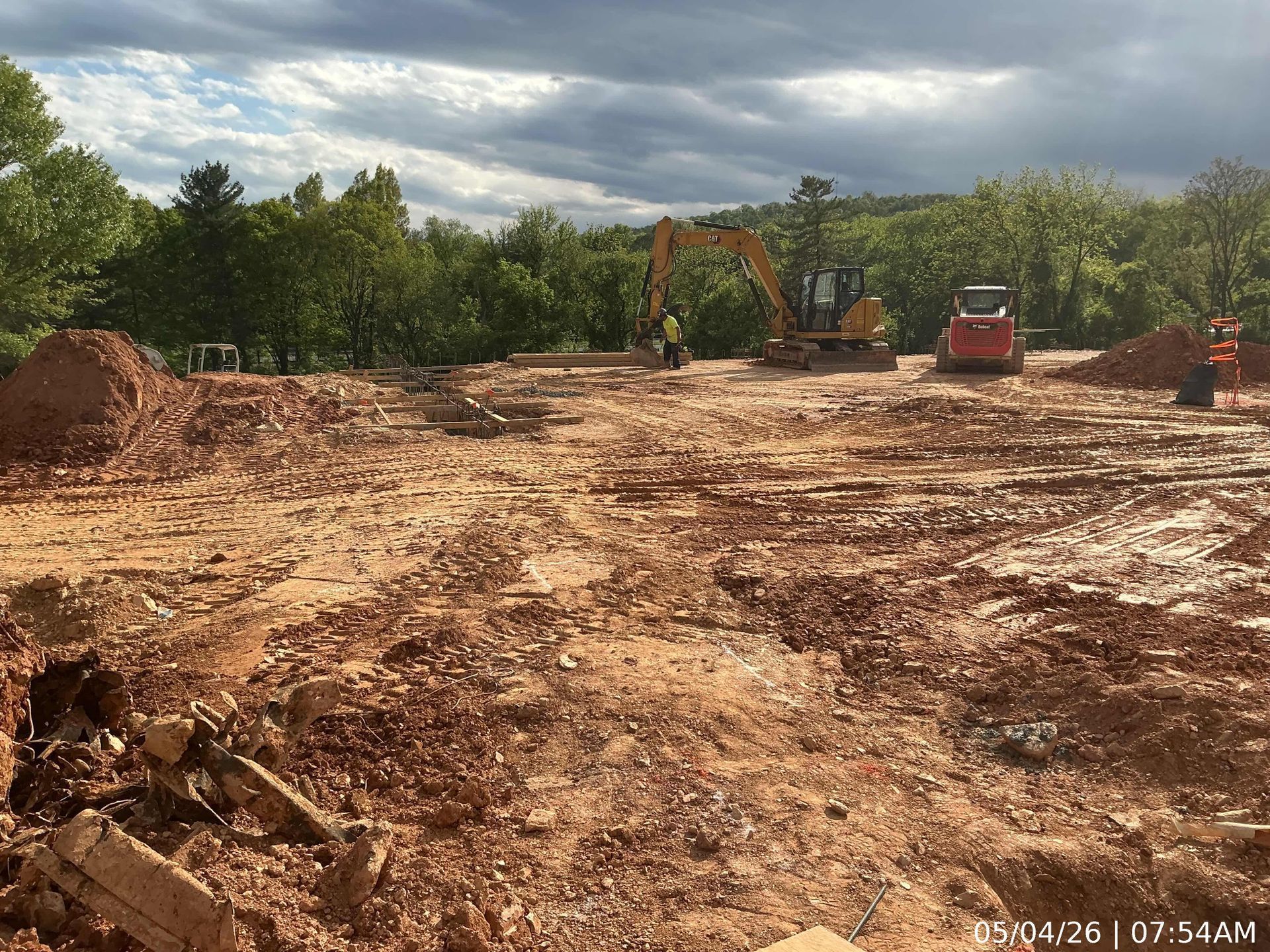 Cleared dirt lot with two excavators and piles of soil under cloudy sky, trees in background