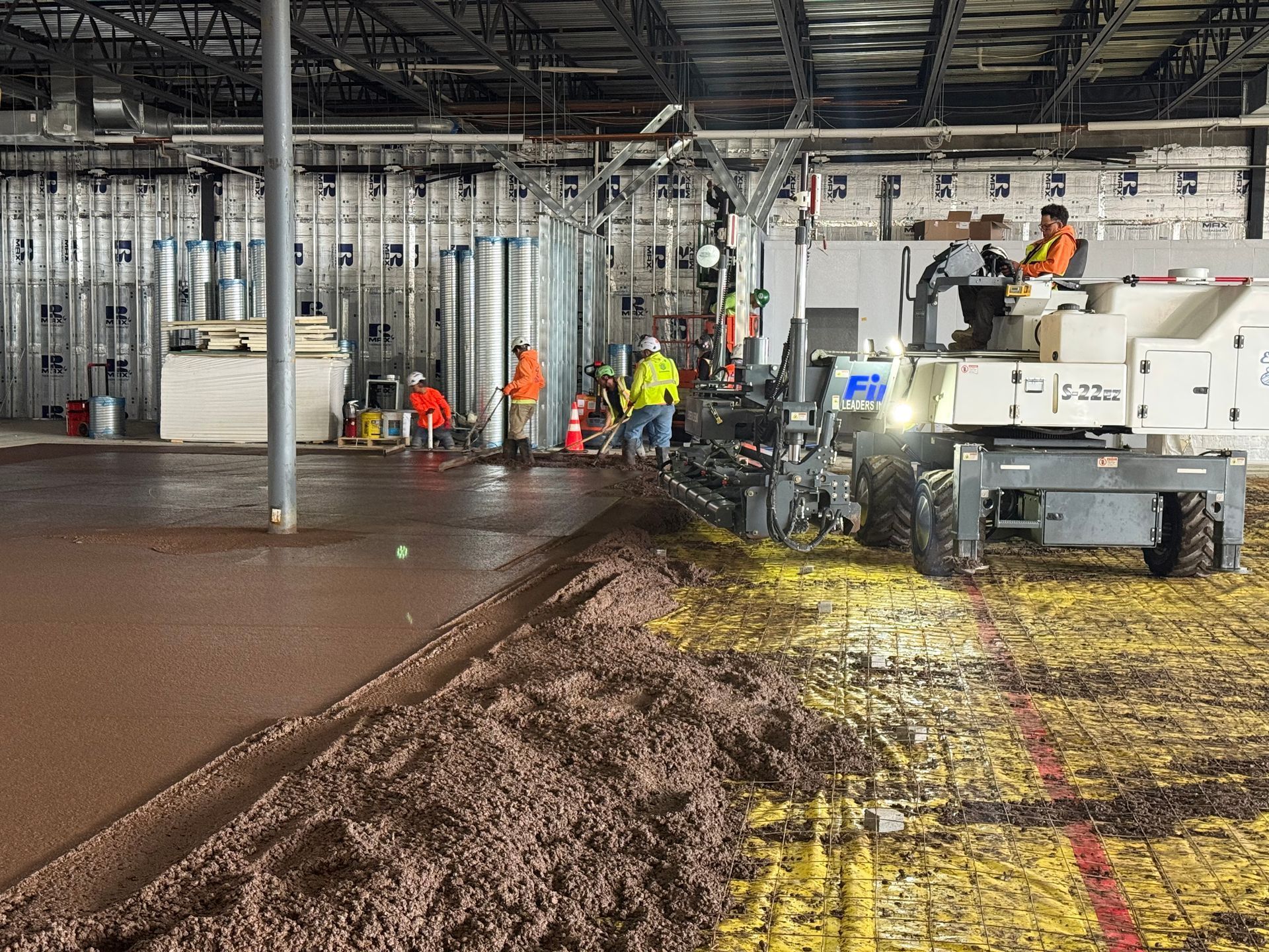 Workers and a paving machine spreading mud on a warehouse floor under construction