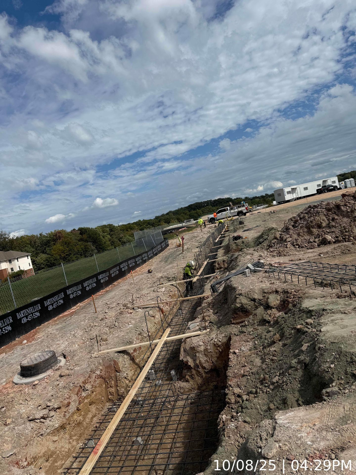 Road construction site with trench, rebar, gravel piles, and cloudy sky