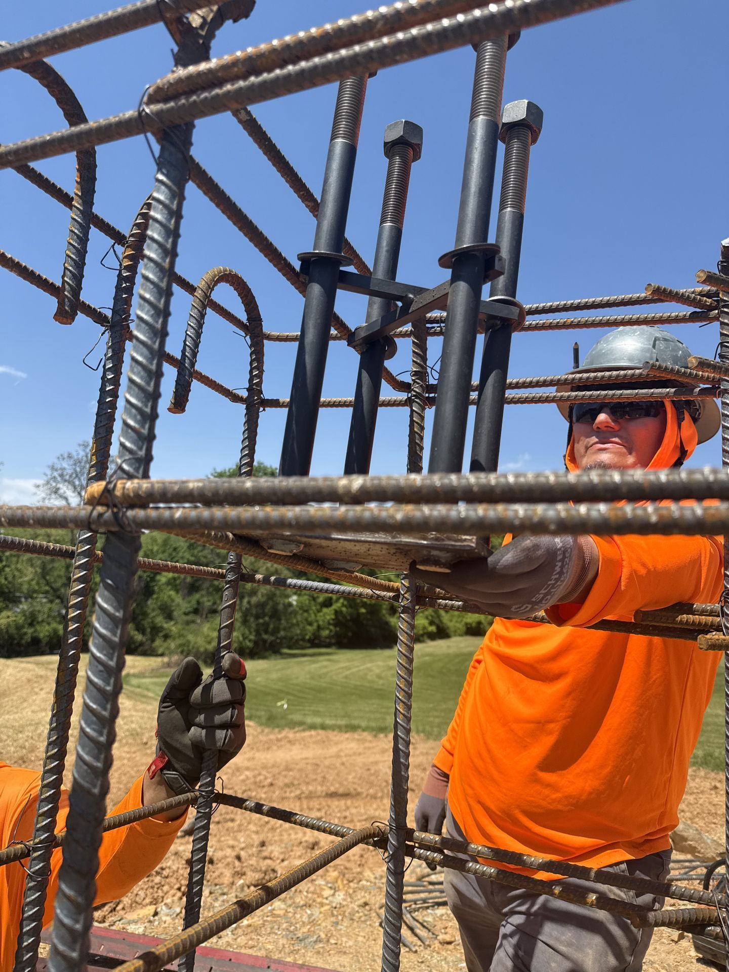 Worker in orange safety gear tying rebar at a construction site outdoors.